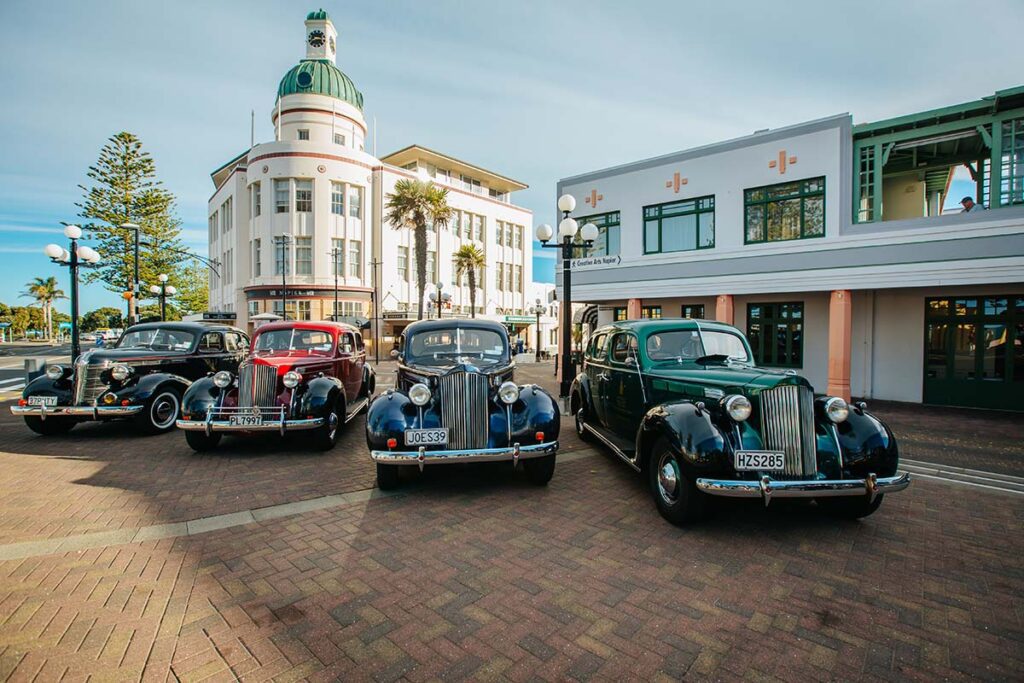 Four vintage cars are parked in a row with an art deco building behind them