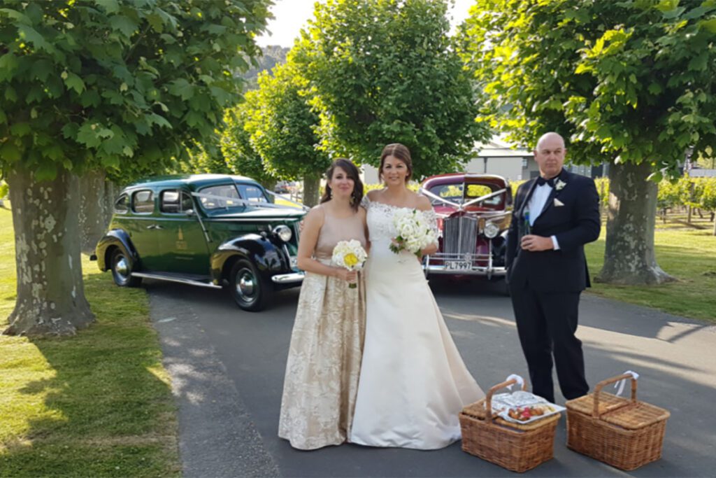 A bride in a white wedding dress, a bridesmaid and man in a tuxedo pose in front of a vintage car